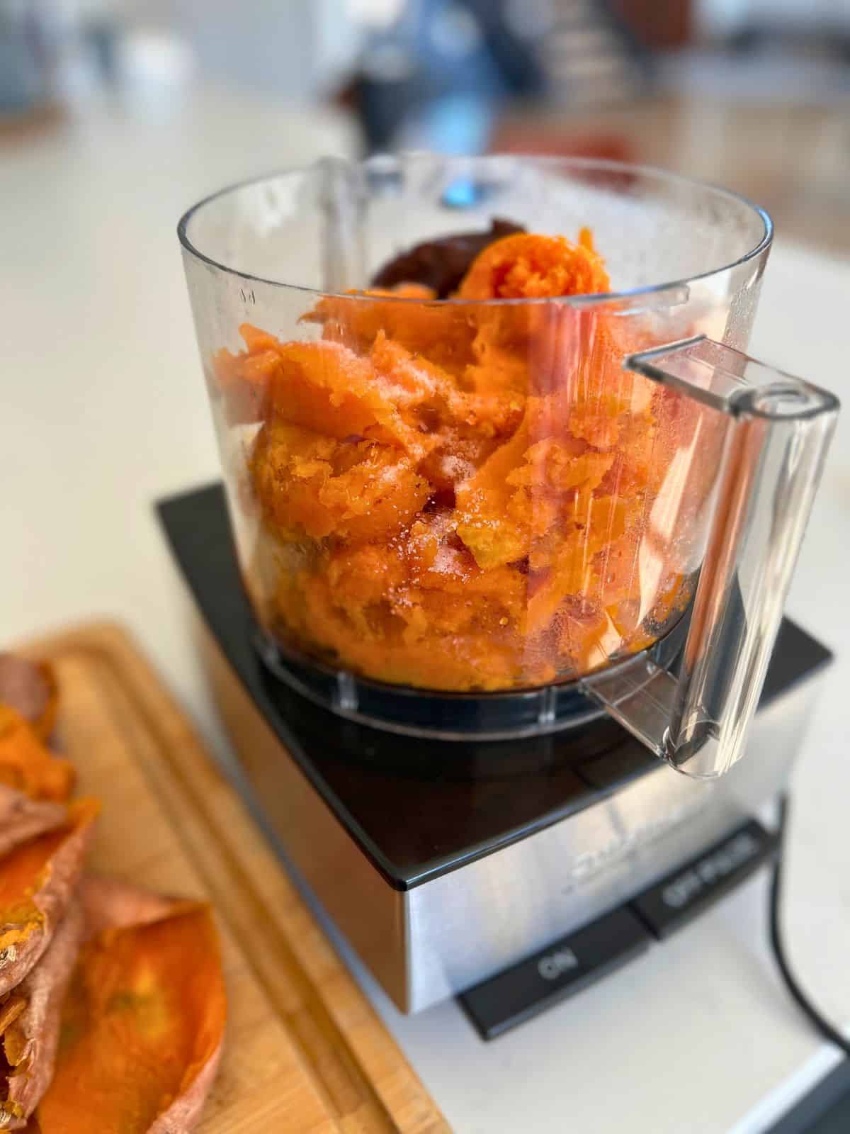 Food processor with scoops of sweet potato pulp, salt, lemon juice, and butter getting ready to puree.