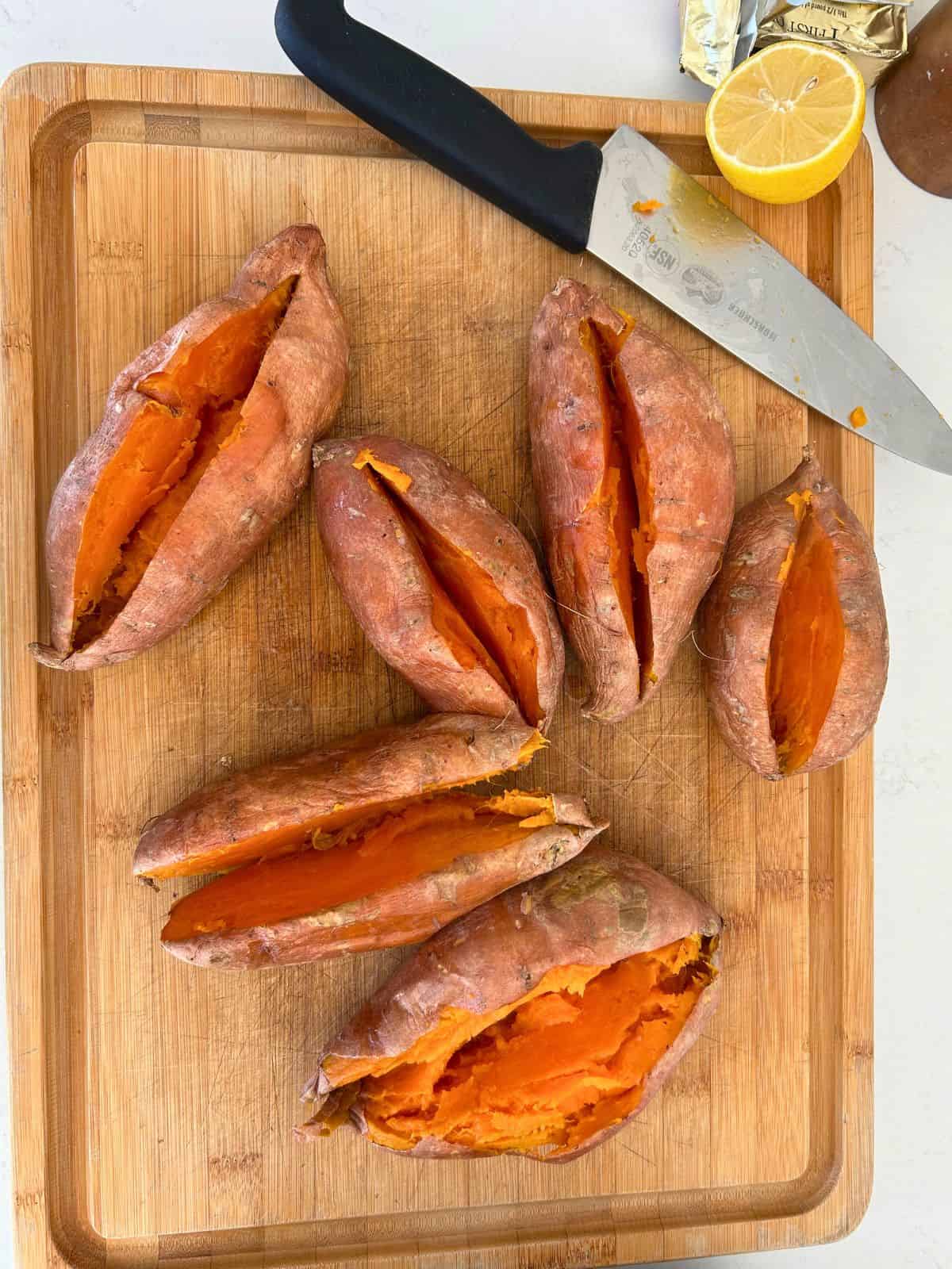 Six whole, cooked sweet potatoes split open on a cutting board with a knife and half a lemon on the corner of the board.