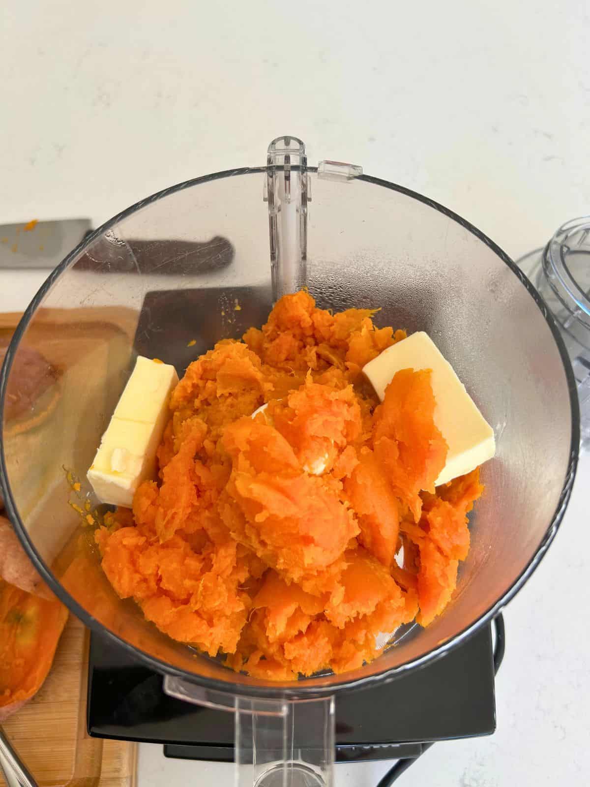 A food processor bowl with part of the cooked sweet potato pulp and chunks of butter so the butter can start to melt.