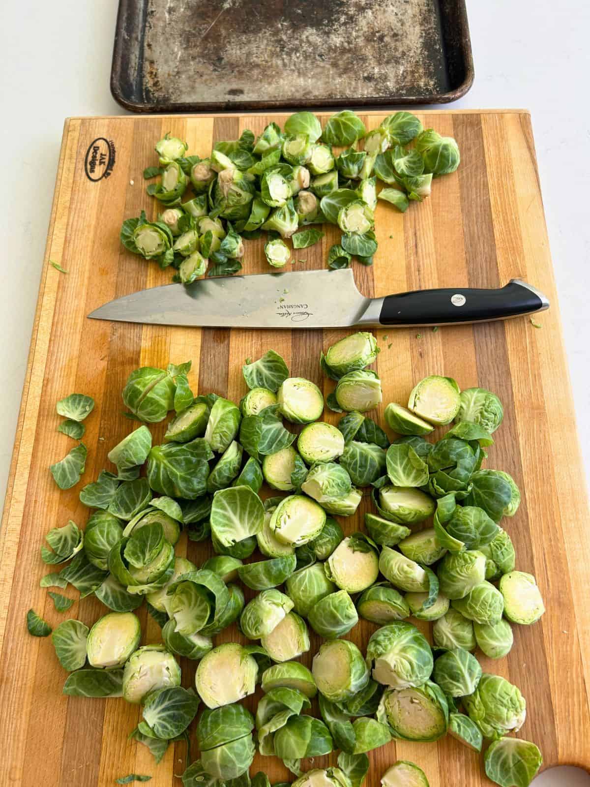 A cutting board containing a pile of brussel sprout scraps, a chefs knife, and larger pile of split brussel sprouts halves.