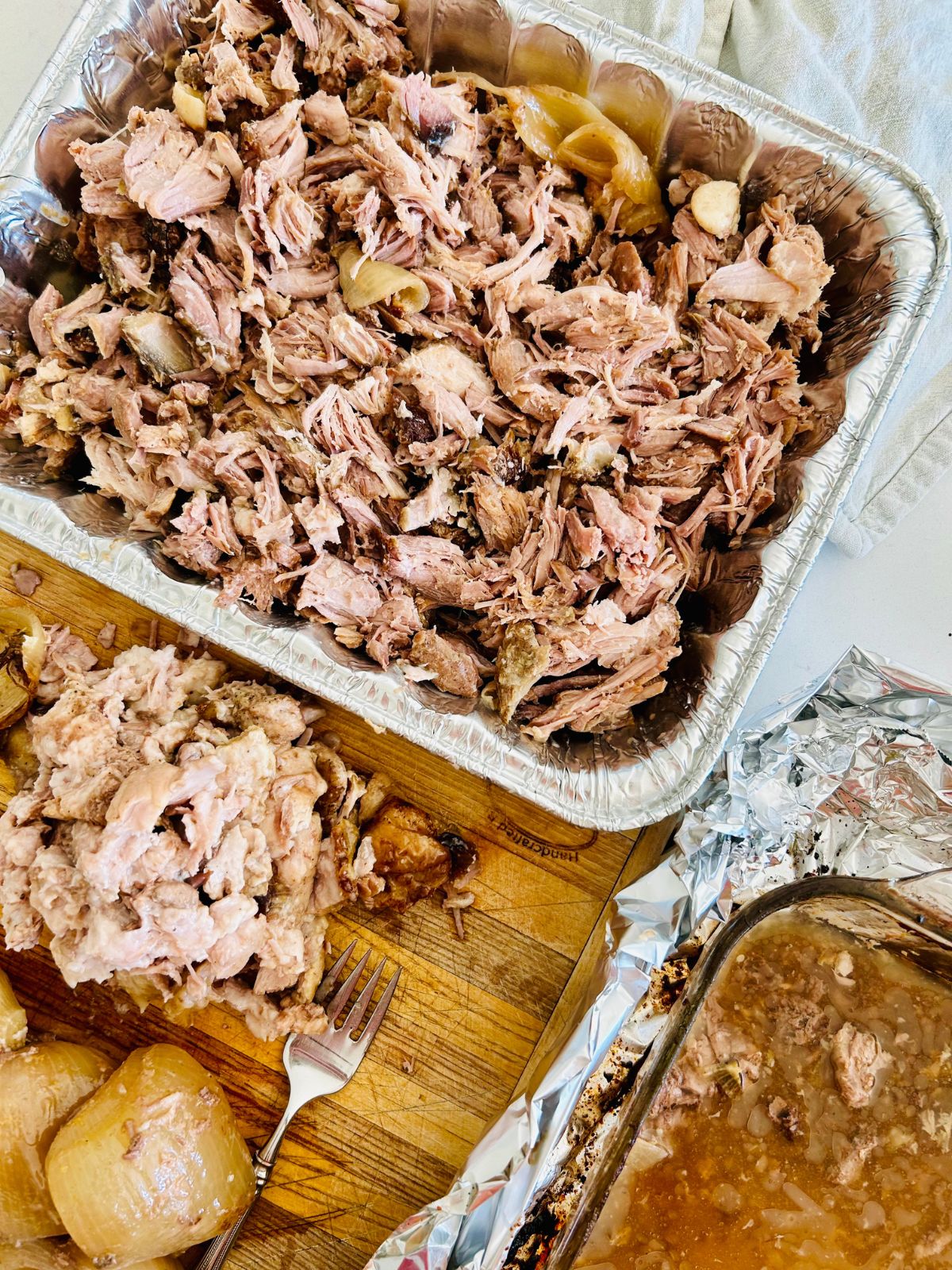 Shredded pork was cooked in a casserole dish and now resting in an aluminum disposable pan next to a cutting board of pork waste.