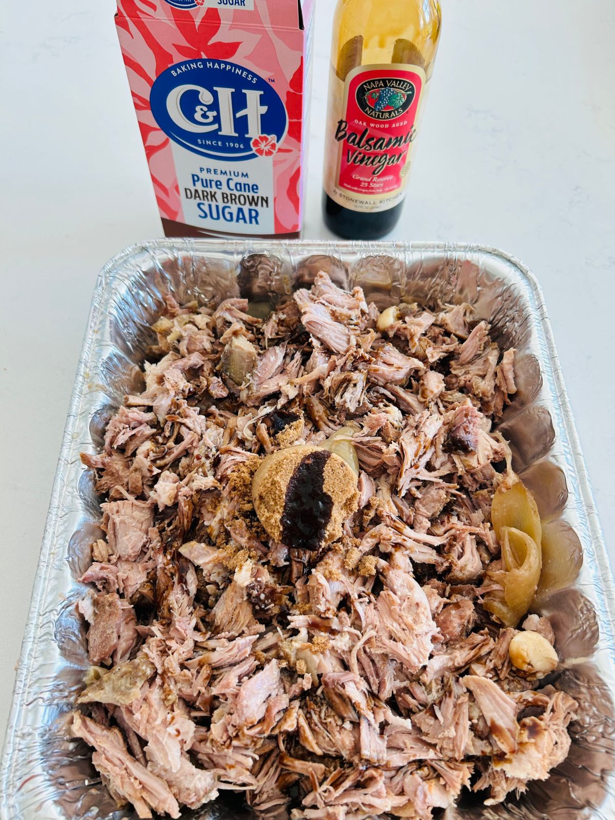 Pulled pork pieces in a disposable aluminum pan with brown sugar and balsamic vinegar on top.