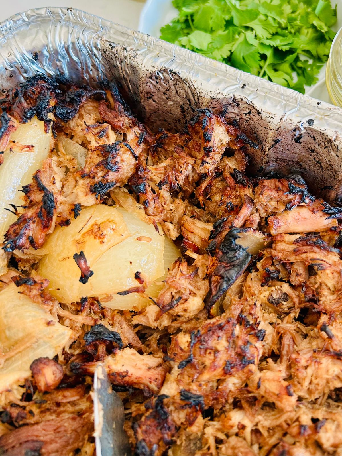 Crispy shredded pork and onions resting in an alumimum disposable pan next to a bowl of cilantro leaves.