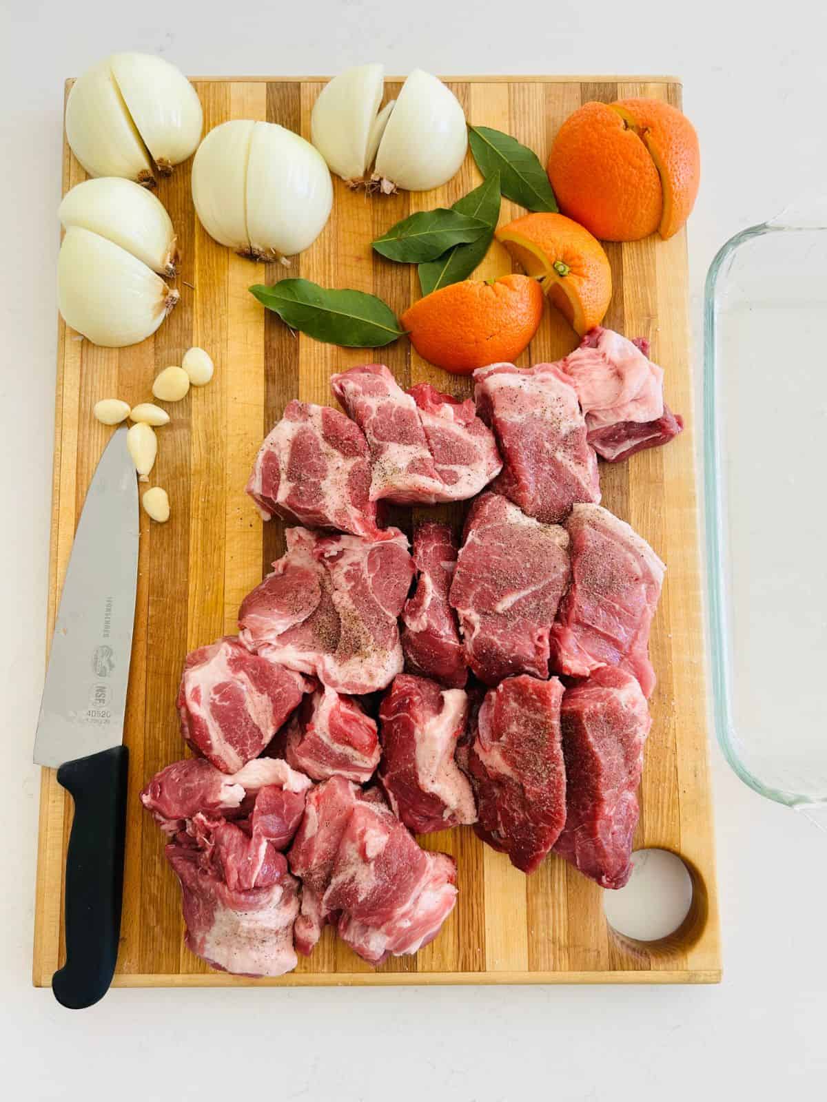 Seasoned pork cubes, quartered onions and orange, bay leaves, and garlic next to a chefs knife on a cutting board.