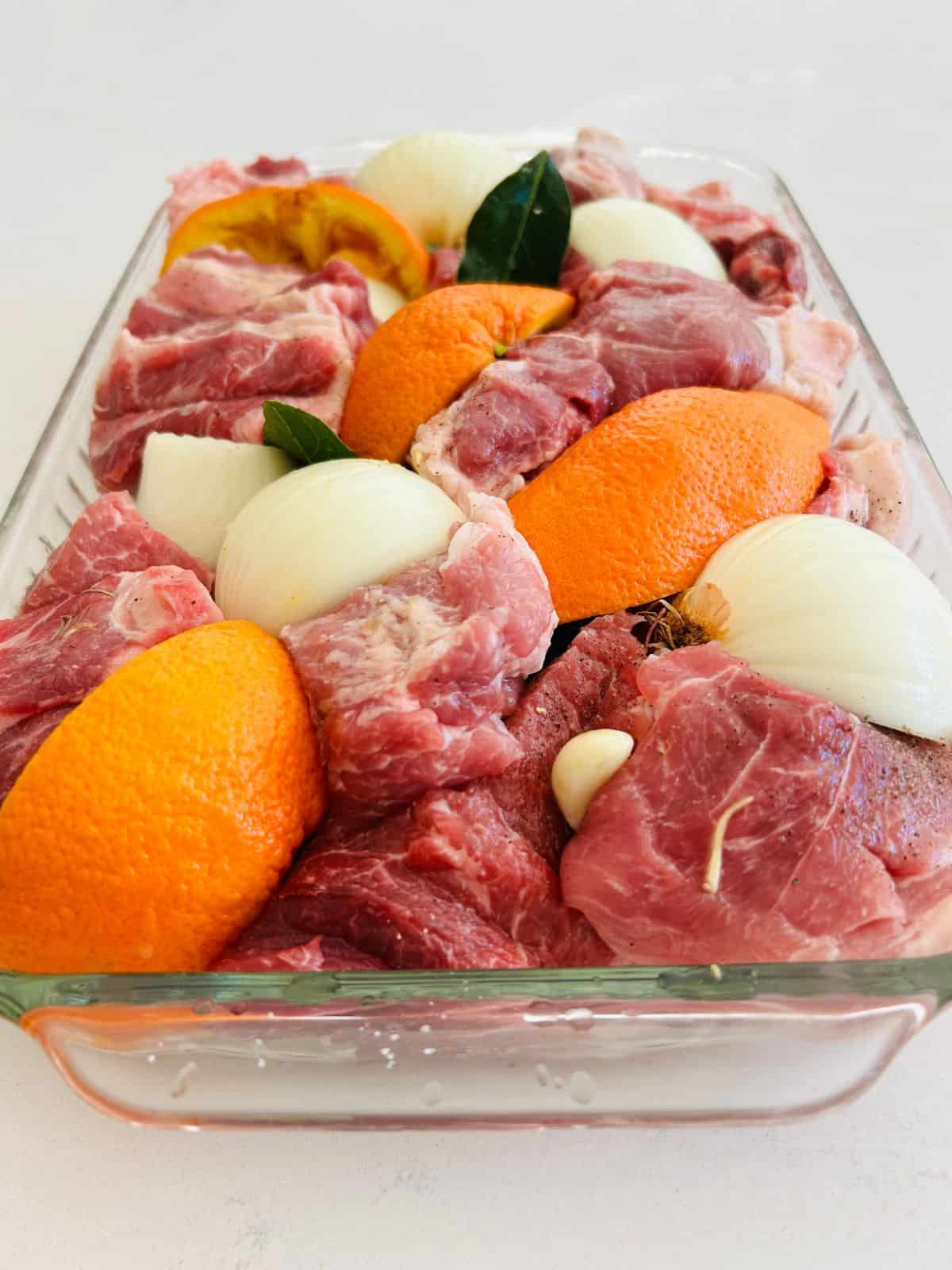 Pork chunks, onion and orange quarters, garlic, and bay leaves nestled together in a glass casserole dish