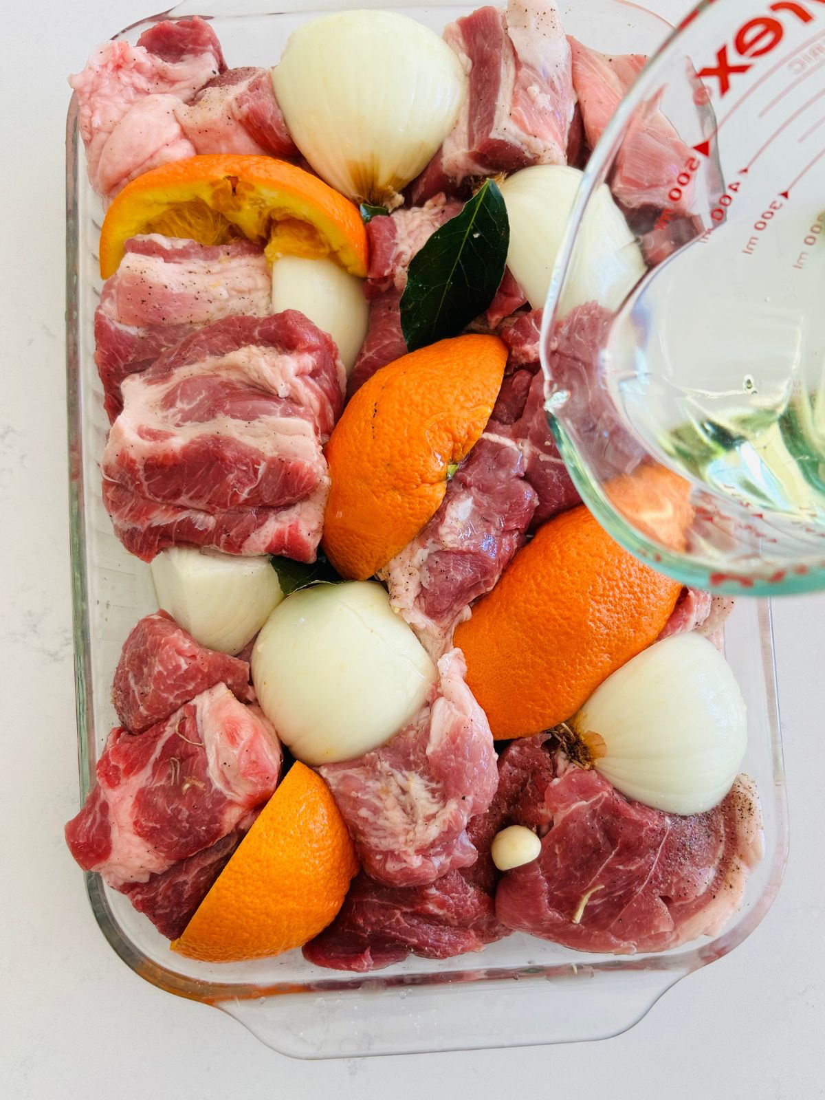 Canola oil is poured over the pork cubes and the ingredients to be roasted in the pyrex dish.