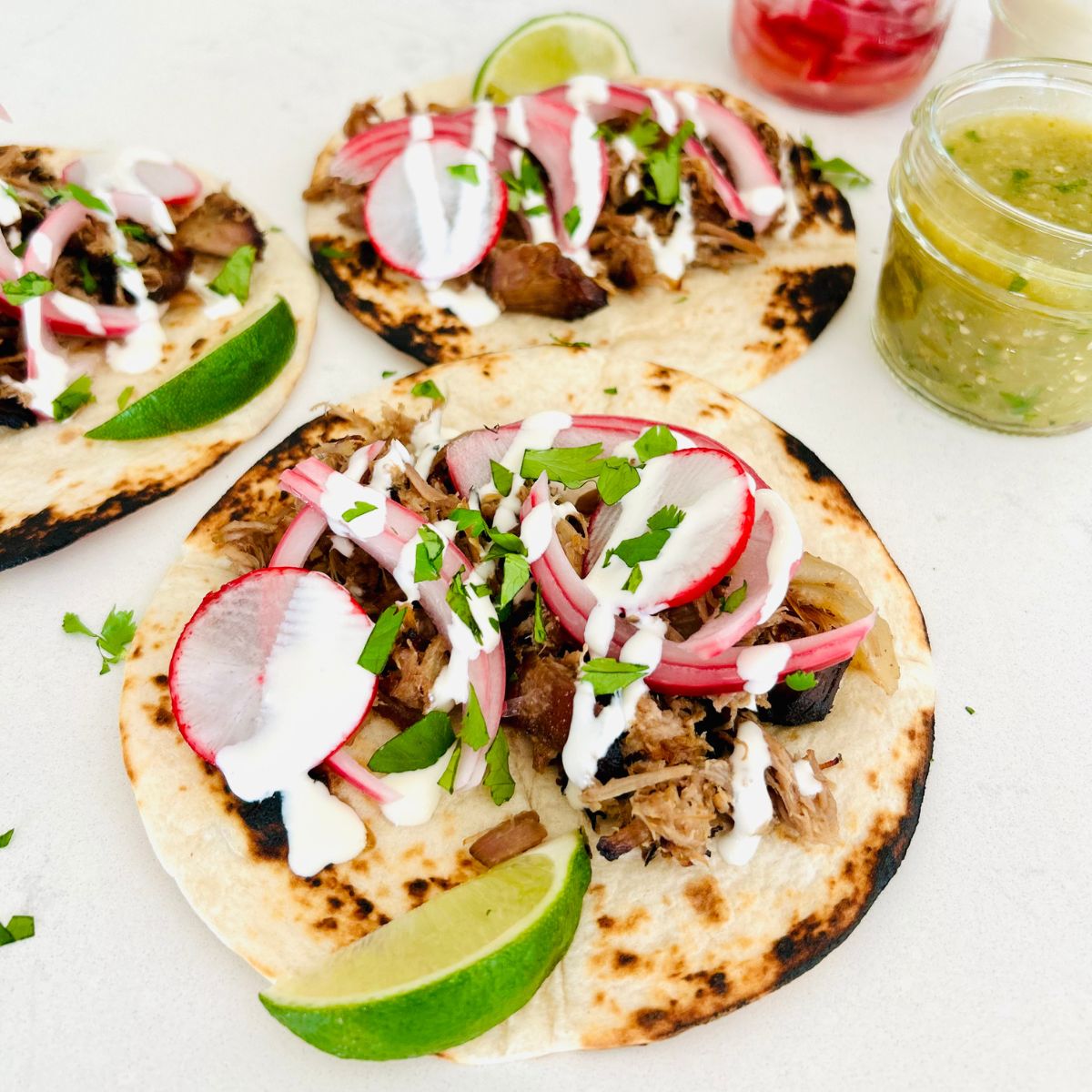 Three flour tortillas with pulled pork, pickled red onions, radishes, crema Mexicana, cilantro and a lime wedge.