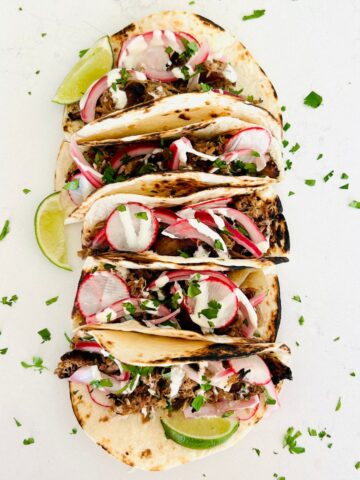 Five small flour tortillas in a row filled with pulled pork, cilantro, radish, pickled red onions, and crema Mexicana.