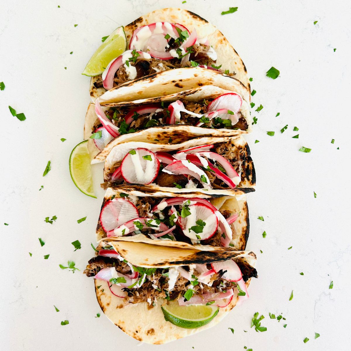 Five small flour tortillas in a row filled with pulled pork, cilantro, radish, pickled red onions, and crema Mexicana.