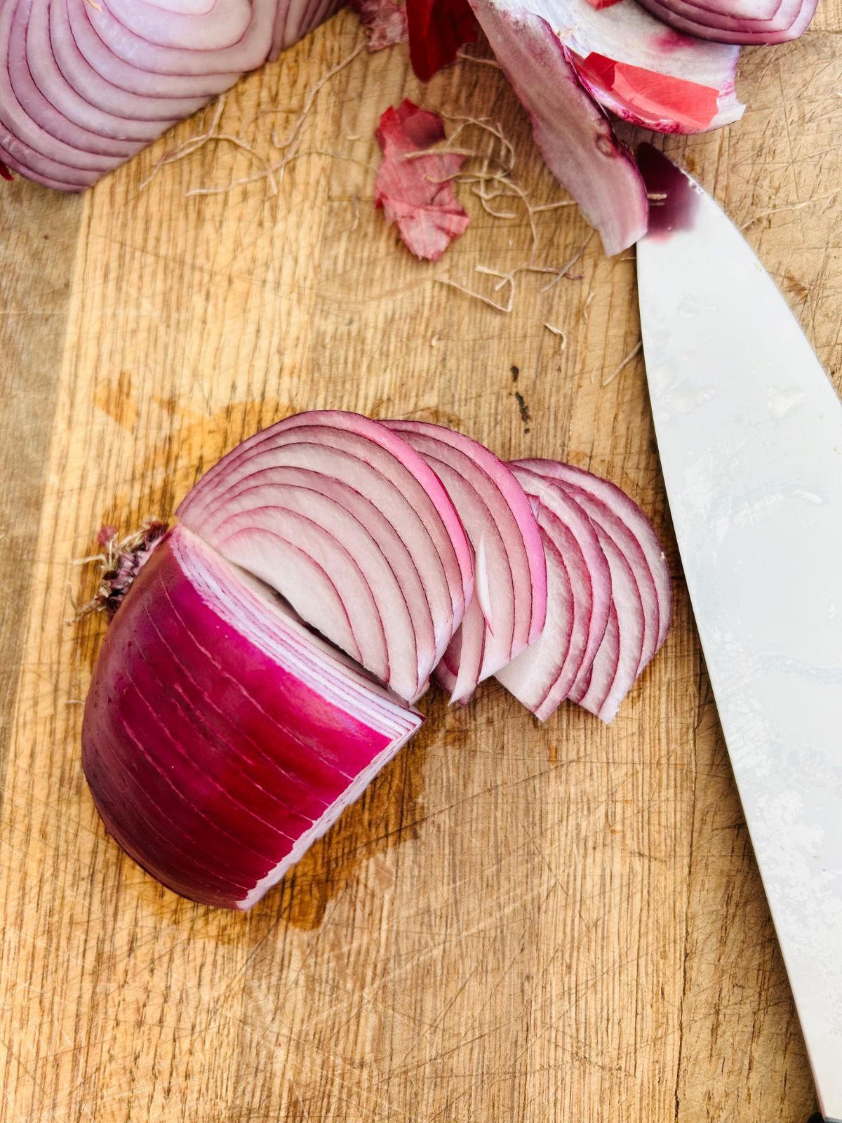 Half of a red onion, thinly sliced and cut wide down on a cutting board next to a chefs knife.