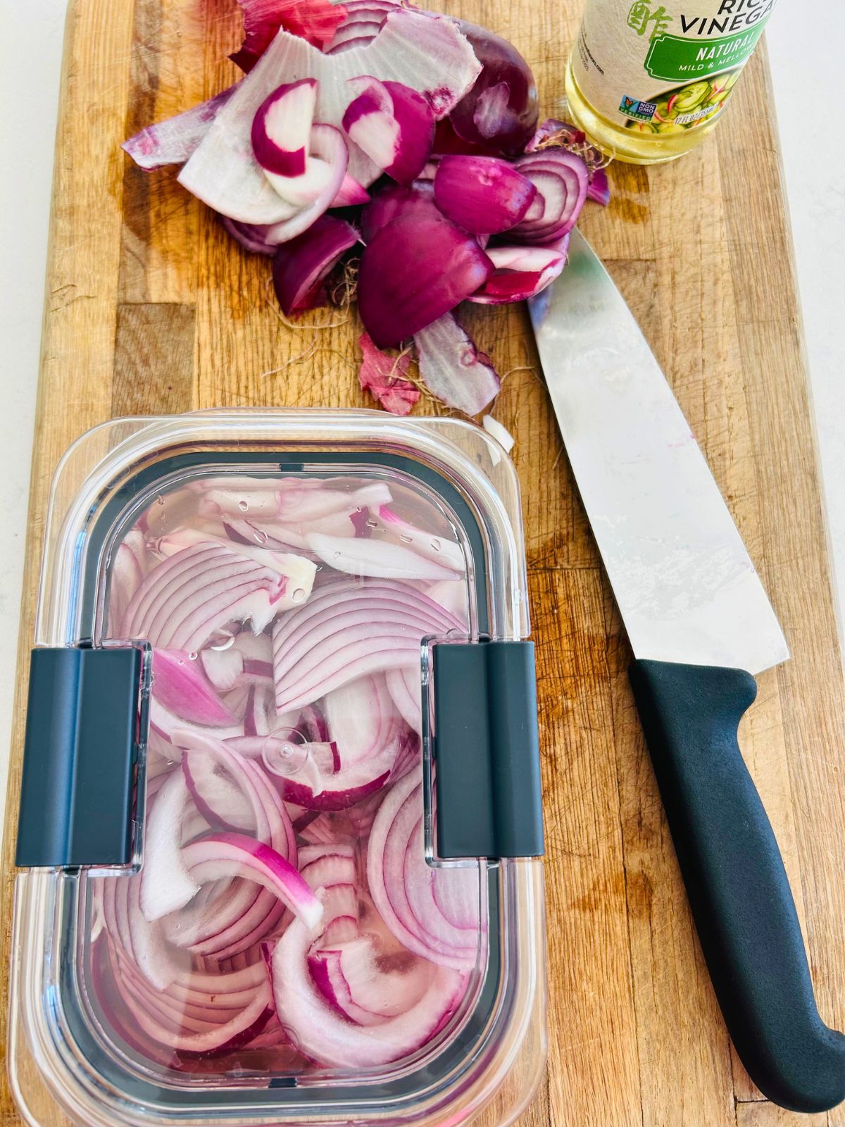 Sliced onions inside a clear, covered container submerged in rice vinegar next to chefs knife and red onion scraps.