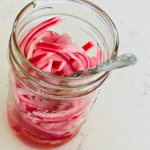 Sliced, pink, pickled red onion pieces resting in a small mason jar with a fork in it.