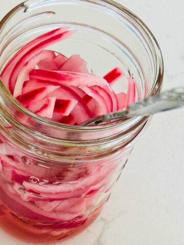 Sliced, pink, pickled red onion pieces resting in a small mason jar with a fork in it.