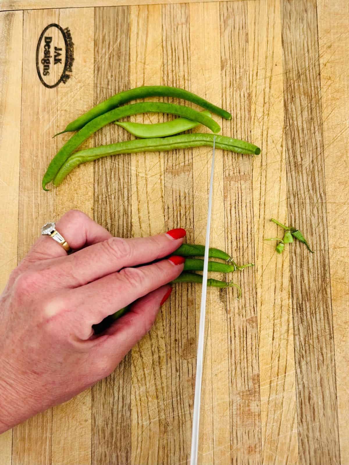 French green beans on a cutting board getting trimmed with a chefs knife.