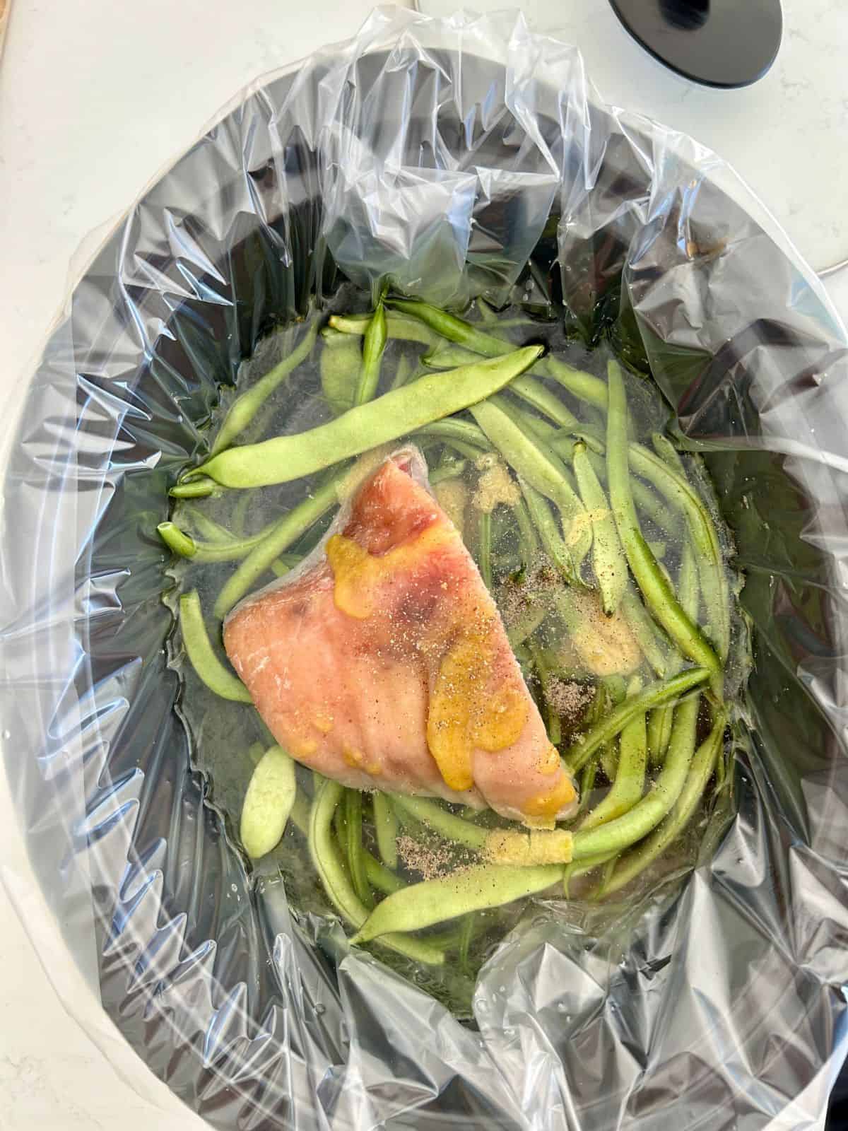 Bright green beans, ham hock, water, bouillon paste, hot sauce, salt and pepper nested in a lined crock pot without a lid.