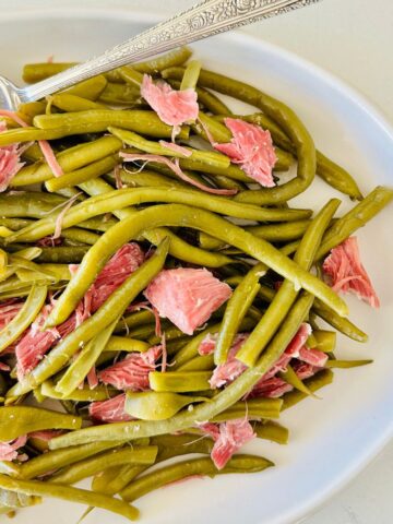 Cooked green beans and pieces of shredded ham on a white plate with a silver serving spoon.