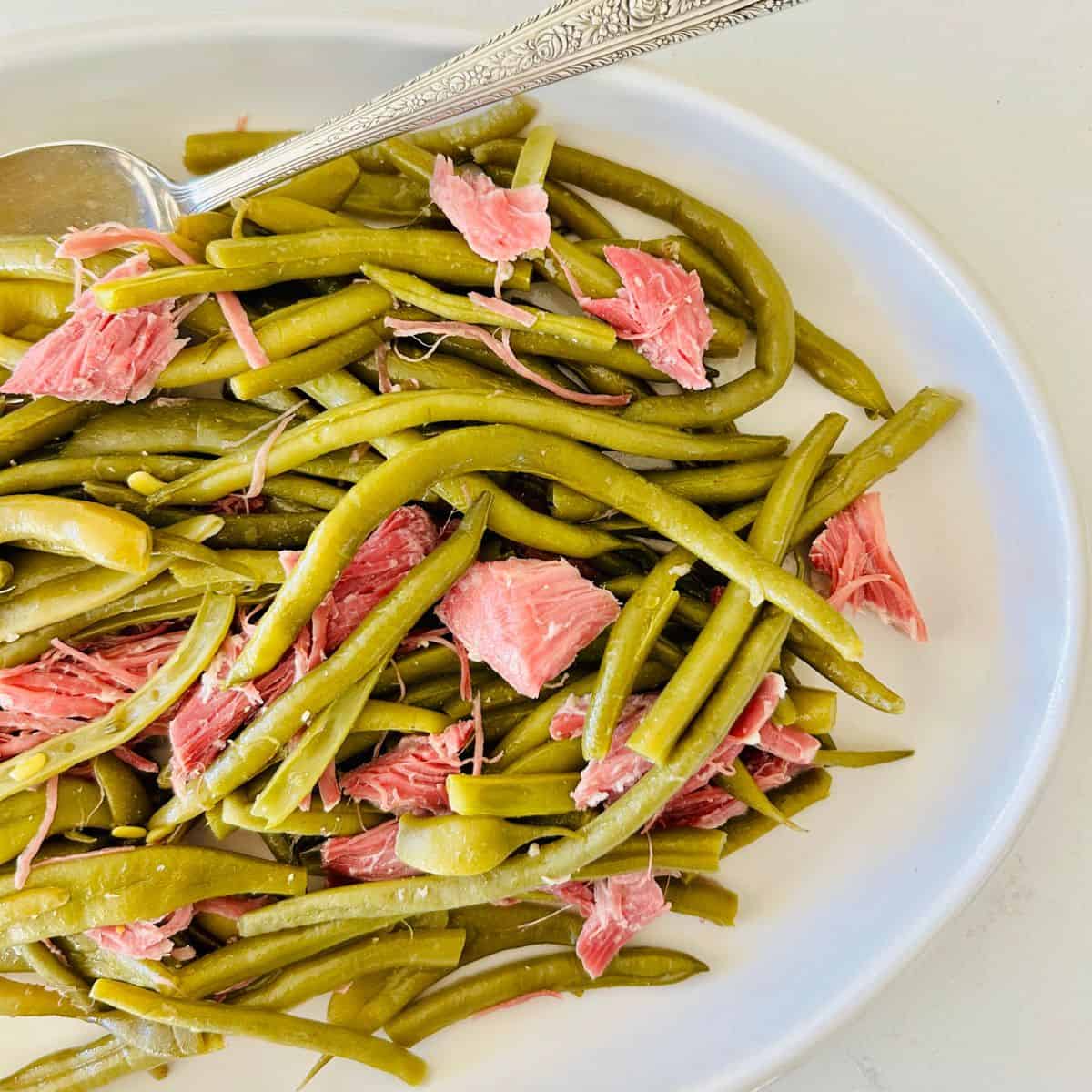 Cooked green beans and pieces of shredded ham on a white plate with a silver serving spoon.