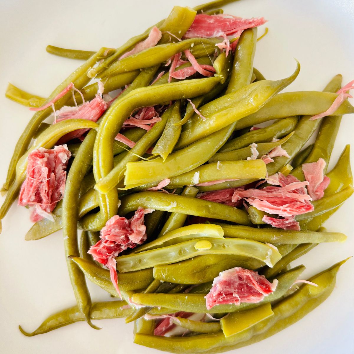 A serving size of cooked and strained green beans interspersed with bite size pieces of ham from the ham shank placed on a white dinner plate.