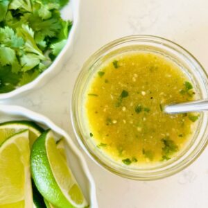 Tomatillo green chili salsa in a mason jar with a spoon next to bowls of lime wedges and cilantro.