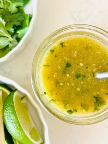 Tomatillo green chili salsa in a mason jar with a spoon next to bowls of lime wedges and cilantro.