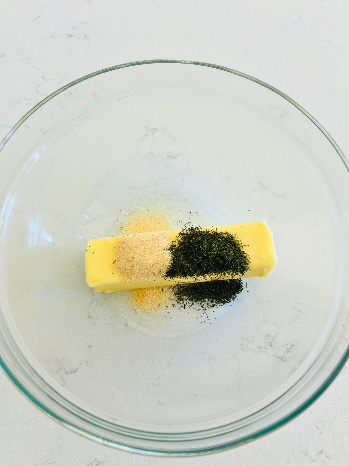 A clear glass bowl with granulated garlic and dried dill on top of a stick of butter.