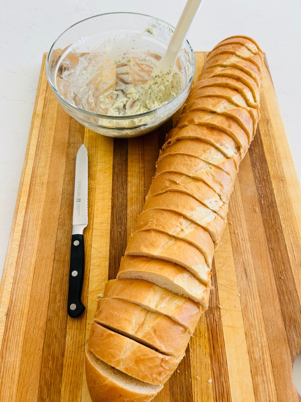 A loaf of french bread that has been sliced into 1 inch thick pieces on a cutting board next to a bread knife and bowl of garlic dill butter.