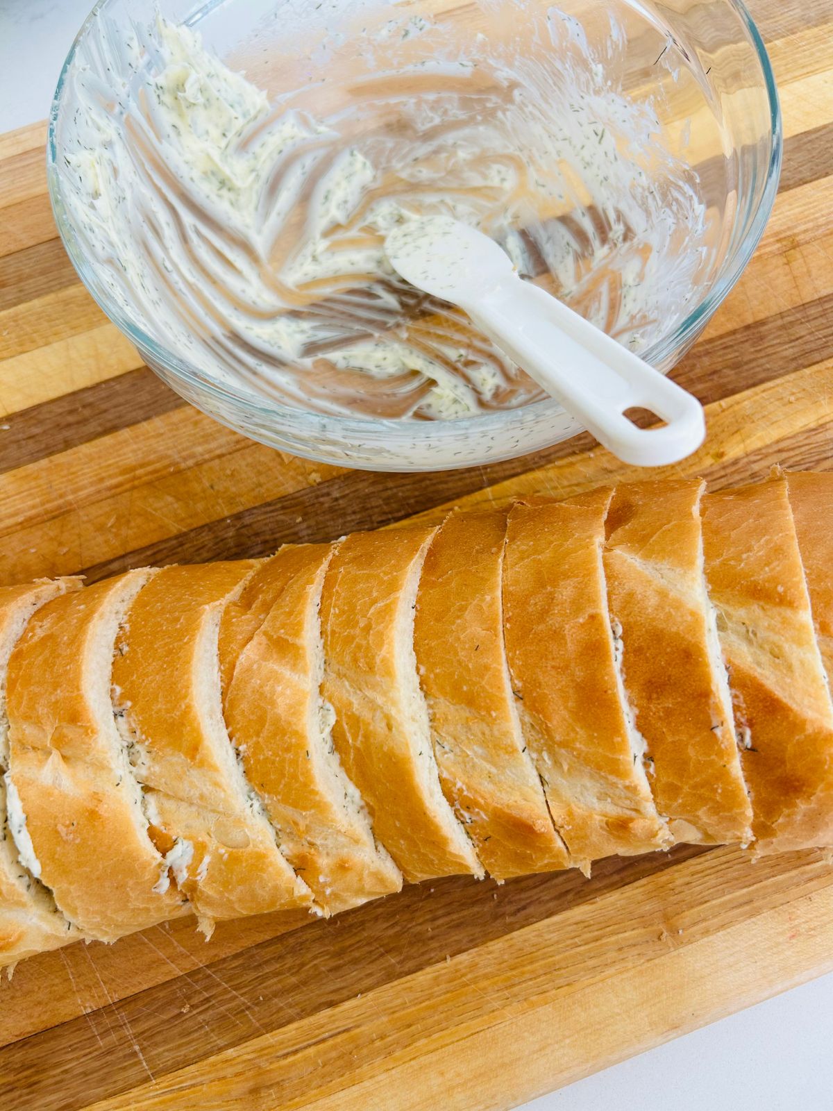 One inch thick slices of buttered french bread laying on a cutting board next to a glass bowl that used to hold the herbed butter.