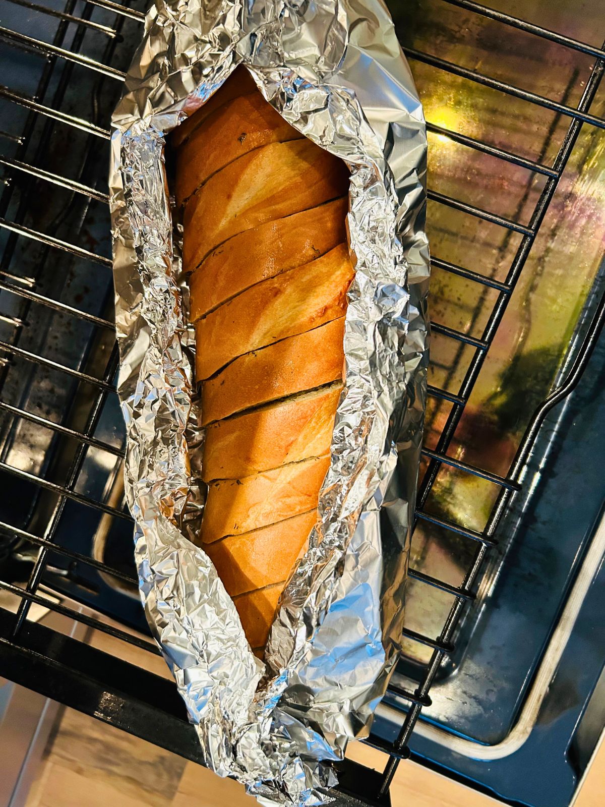 A loaf of garlic bread wrapped in foil that has been partially cracked open on the rack of the oven.