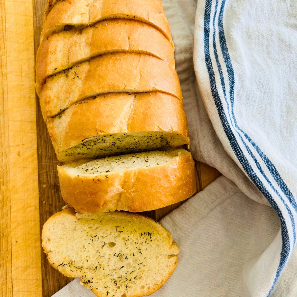 Toasted garlic and dill French bread sliced thickly on a cutting board next to a tea towel.