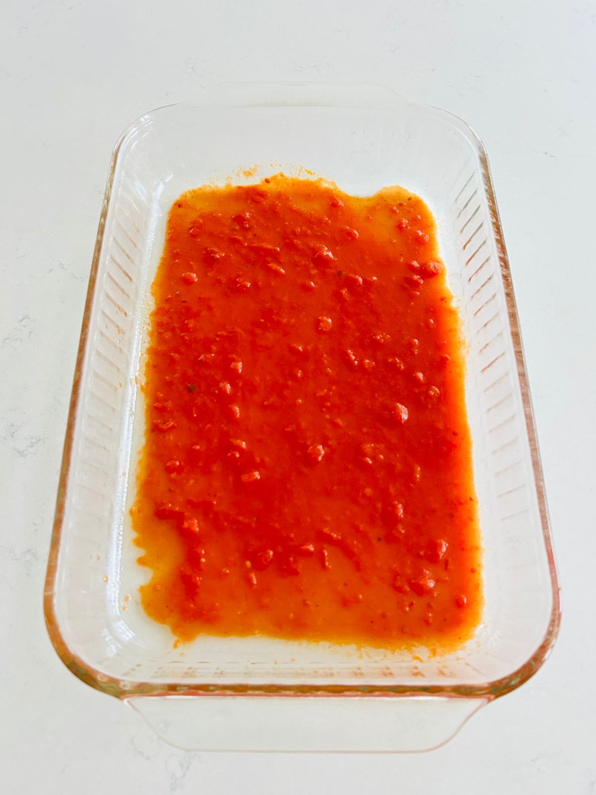 A Pyrex casserole dish with one quarter of a cup of marinara in it for the first step of making a lasagna.