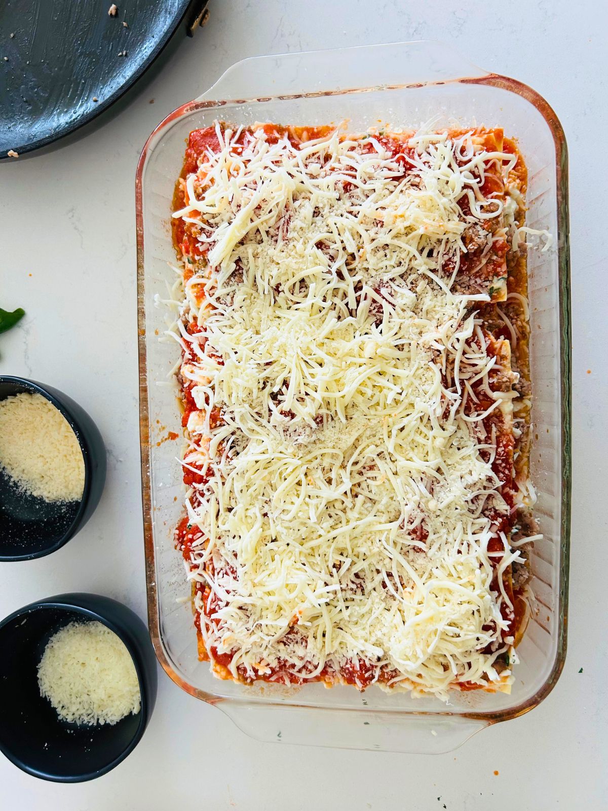 Lasagna that has been layered in a pyrex dish next to bowls of grated Parmigiano Reggiano and Pecorino cheese.