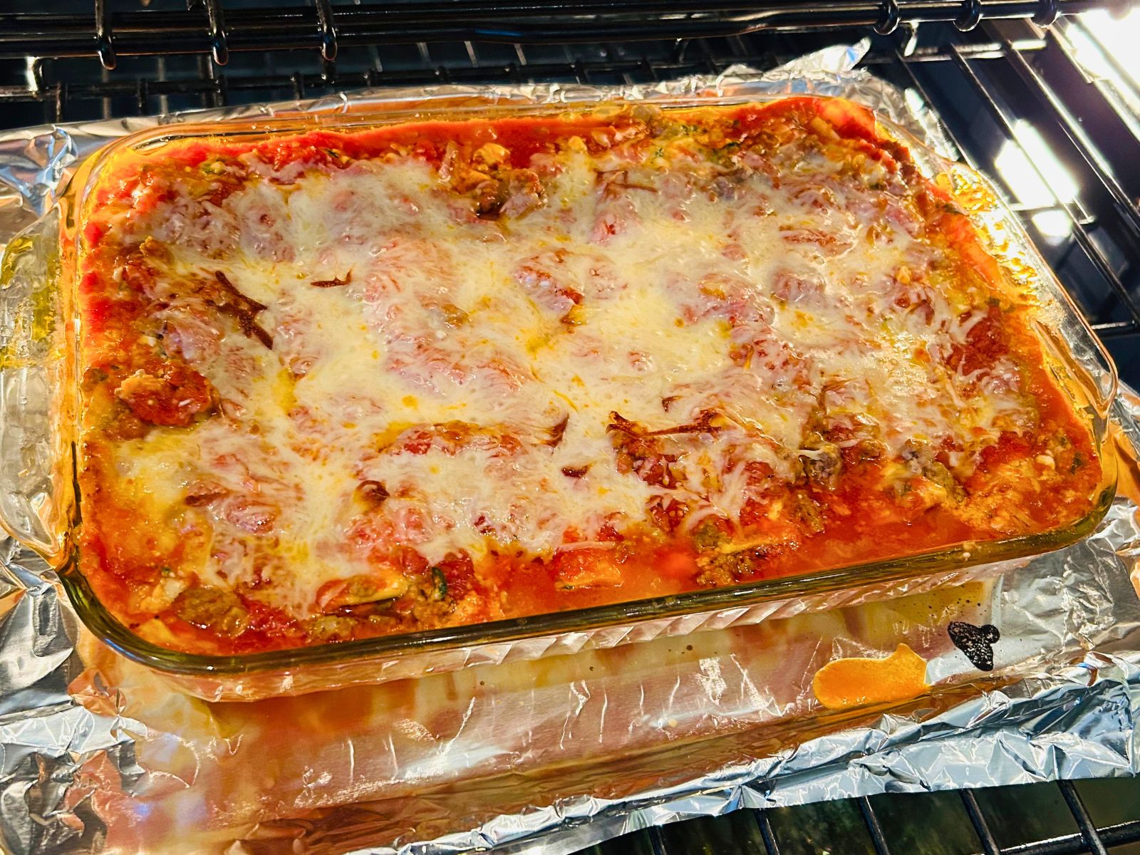 A casserole dish of mostly cooked lasagna on top of a foil lined cookie sheet in the oven.