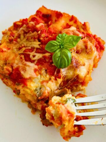 A serving of lasagna on a white plate with a sprig of fresh basil on top with it's corner cut off and held on top of a fork.