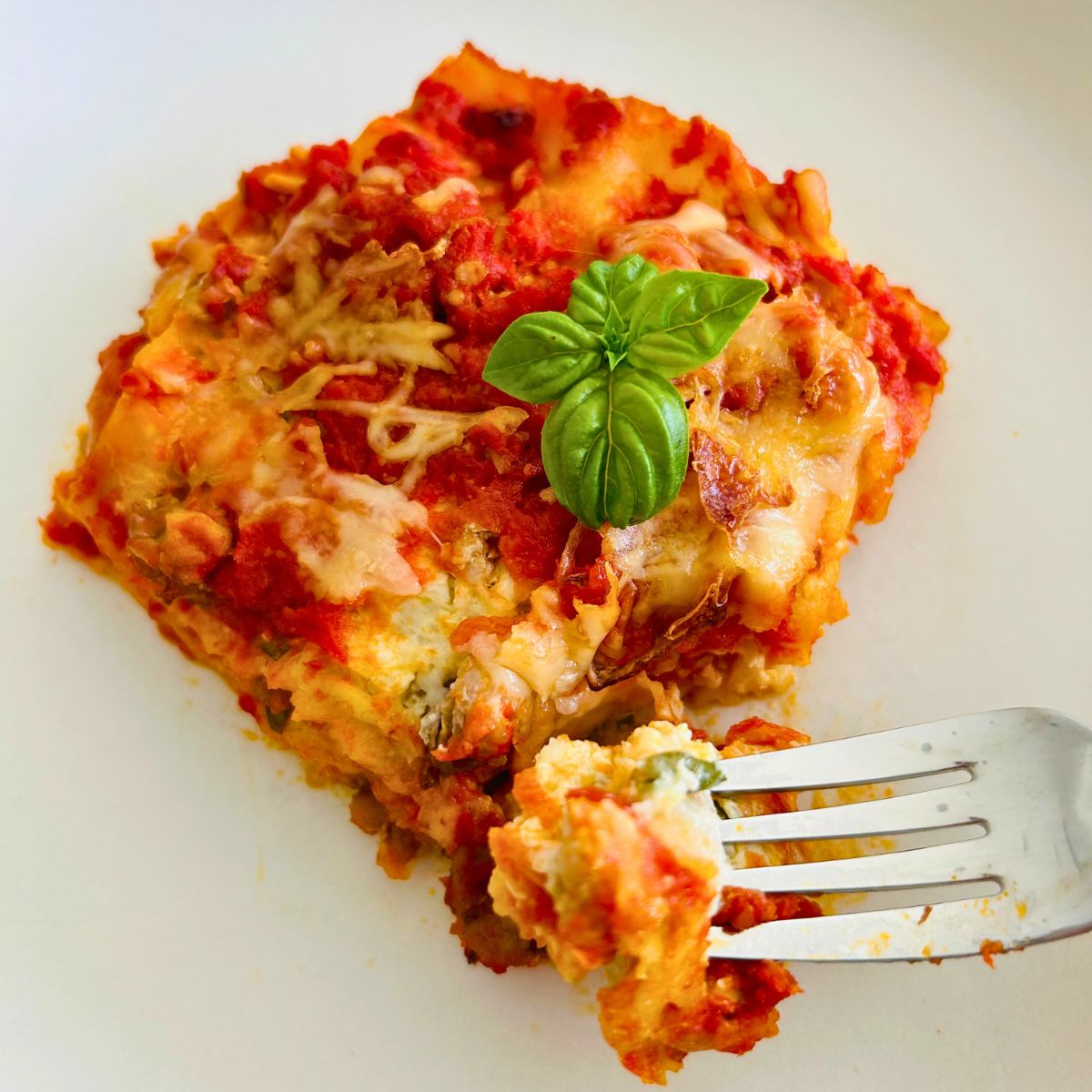 A serving of lasagna on a white plate with a sprig of fresh basil on top with it's corner cut off and held on top of a fork.