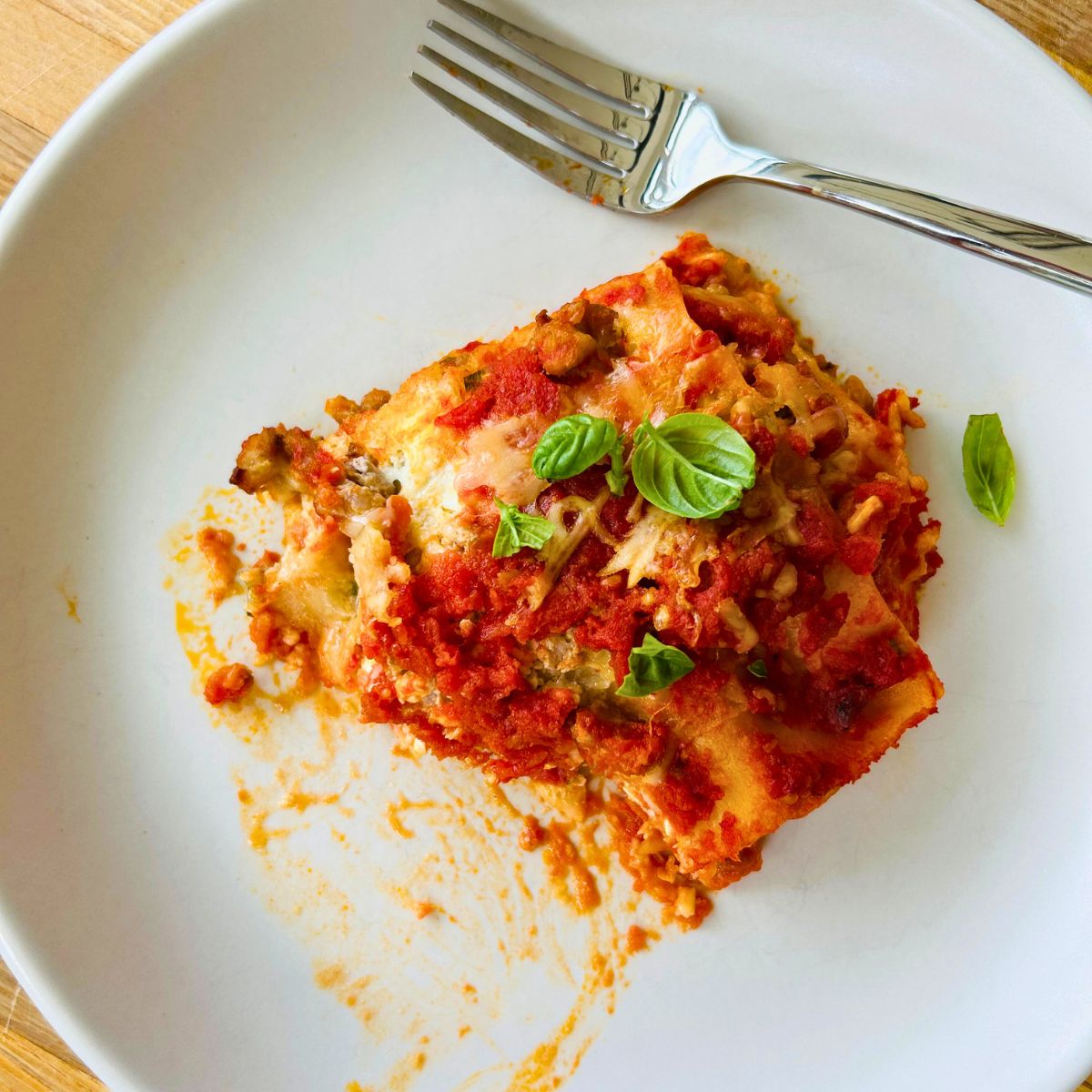 A piece of partially eaten lasagna with a dirty fork on top of a white plate.