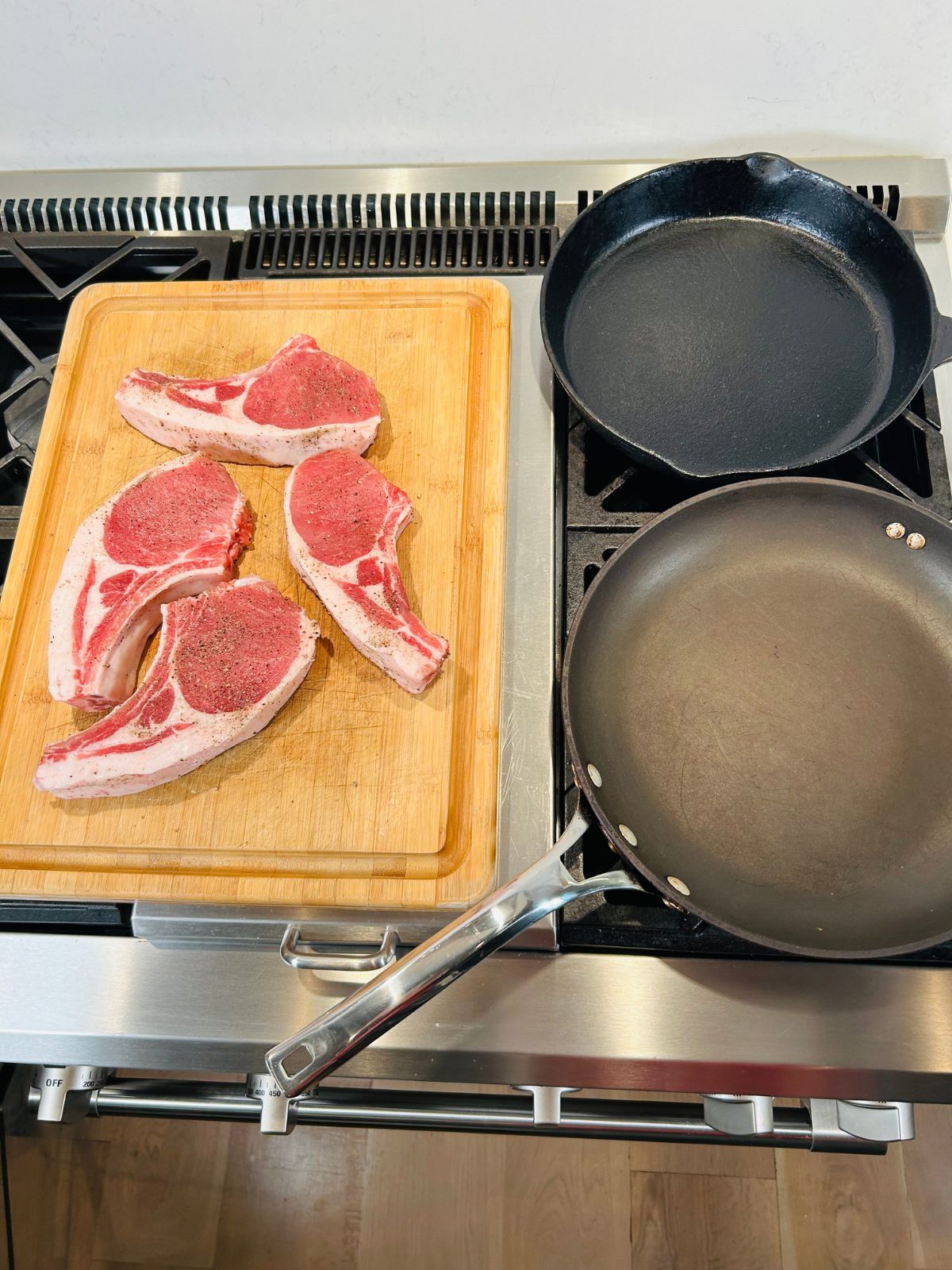 Two oven proof skillets heating up with the four pork chops waiting on a cutting board next to them.