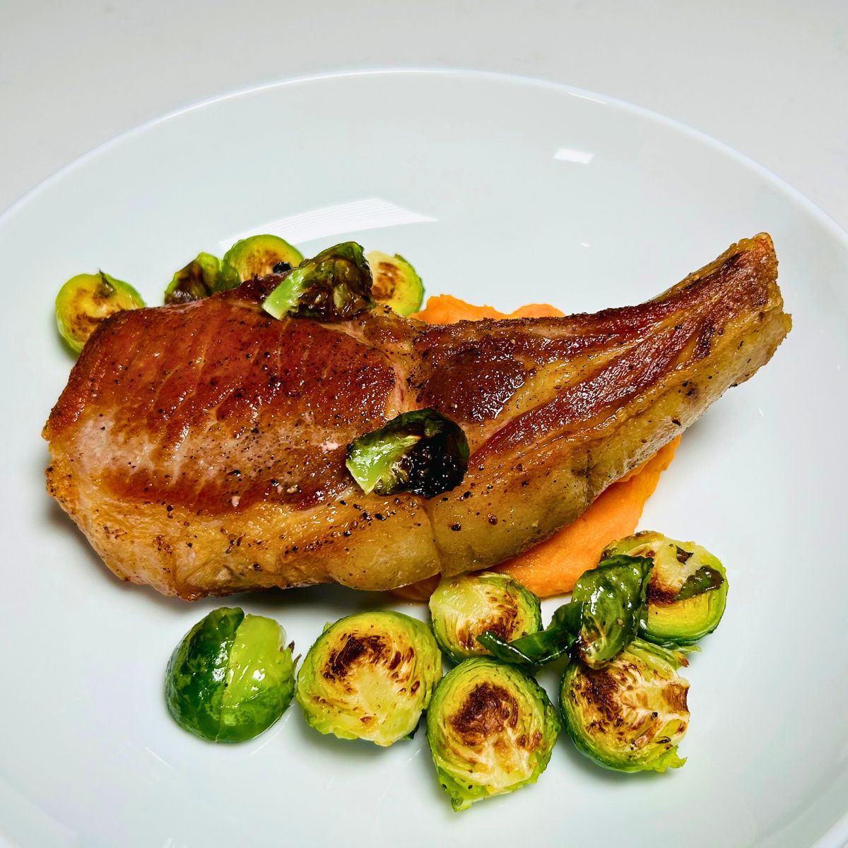 Seared, bone-in pork chop on a white plate with brussel sprouts and sweet potato puree.