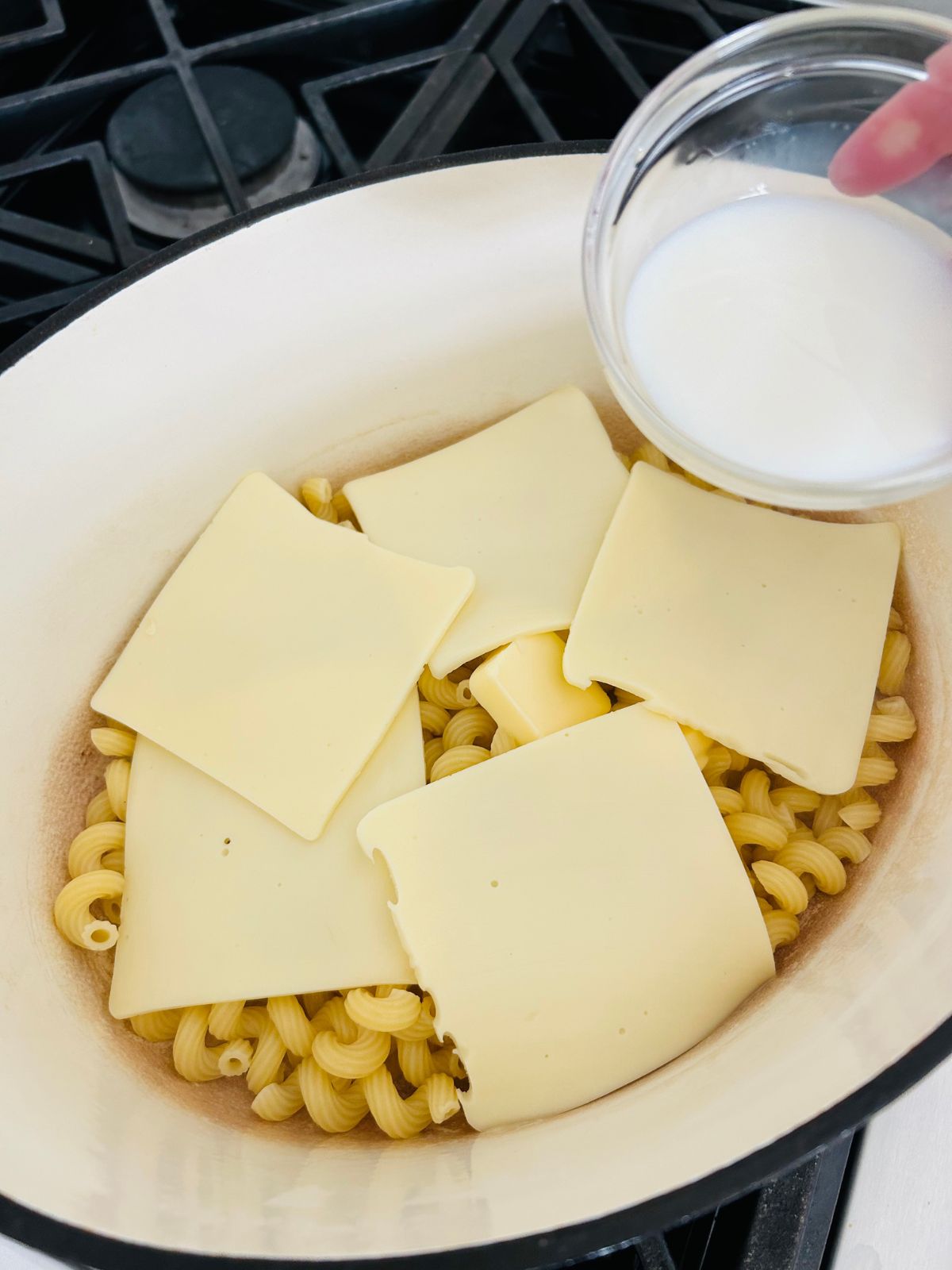 A pot of cooked macaroni pasta with American Cheese slices and butter on top while milk is ready to pour into the pot.