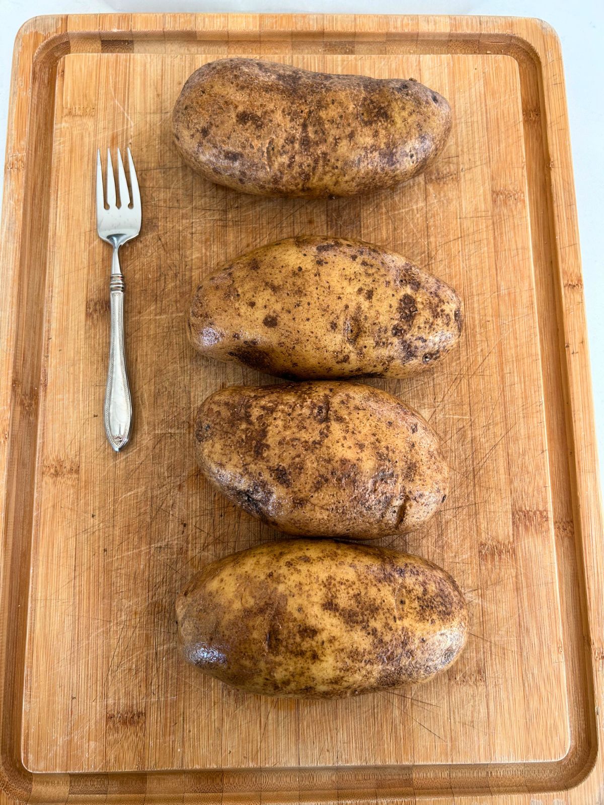 Four washed baked potatoes on a cutting board next to a fork.