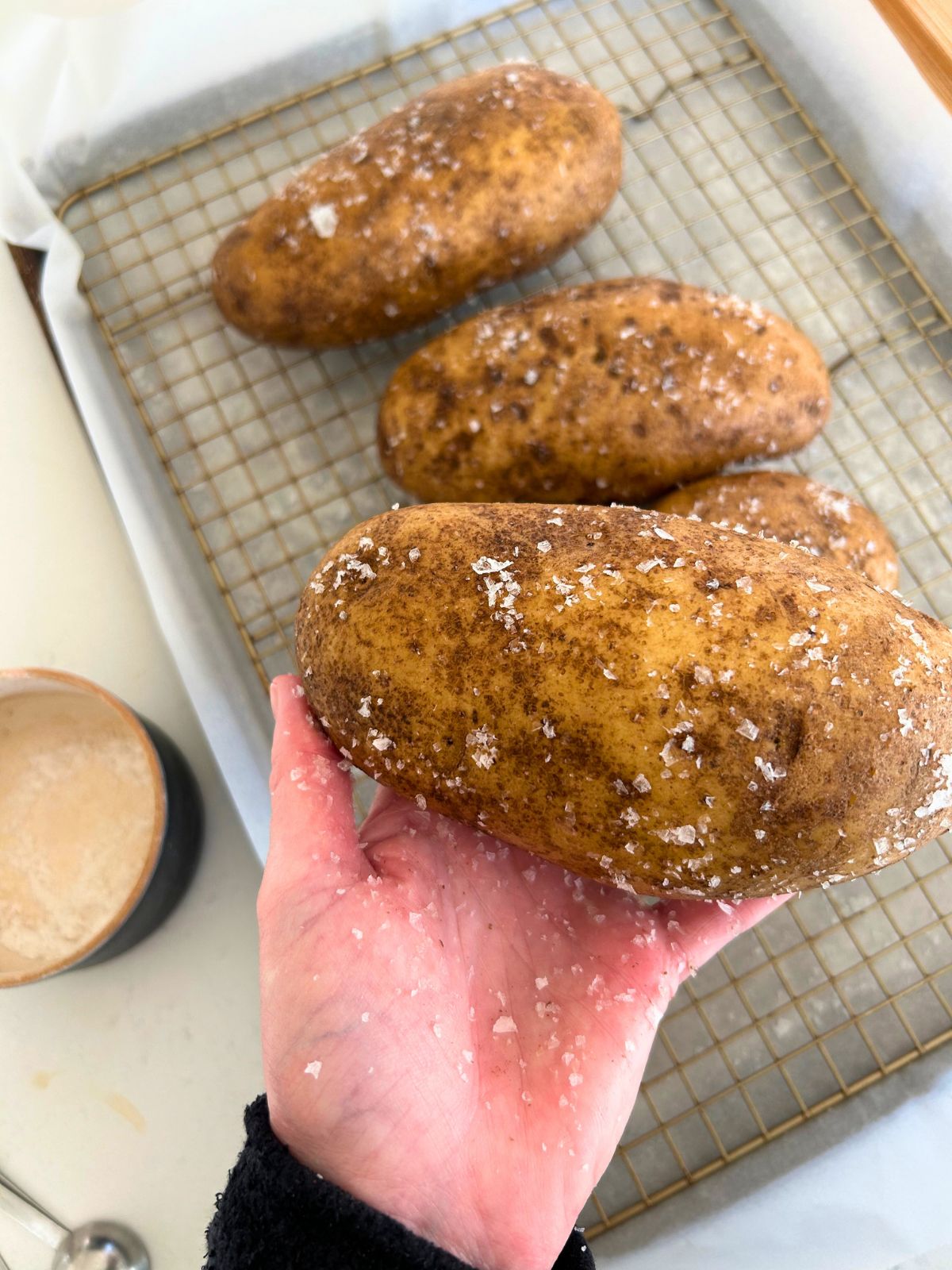 Hand sprinkling Maldon salt on a damp baked potato with a cookie sheet underneath.