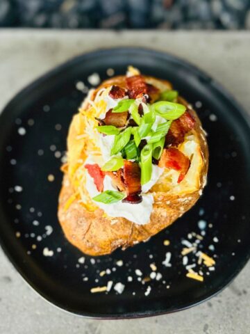 A steakhouse baked potato with toppings of butter, sour cream, bacon, cheese, and green onions served on a black plate.