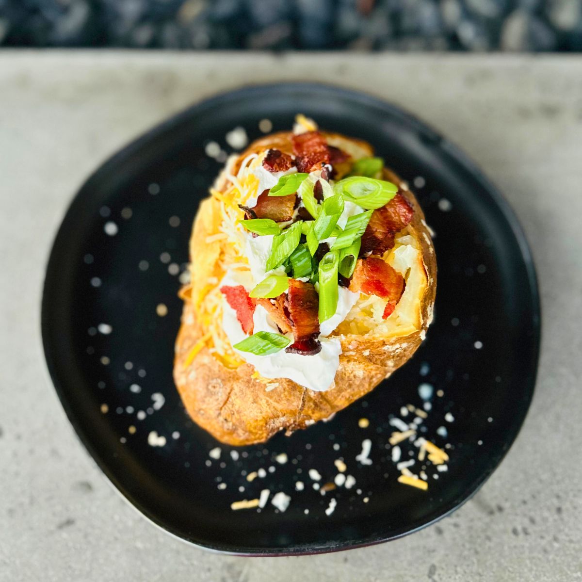 A steakhouse baked potato with toppings of butter, sour cream, bacon, cheese, and green onions served on a black plate.