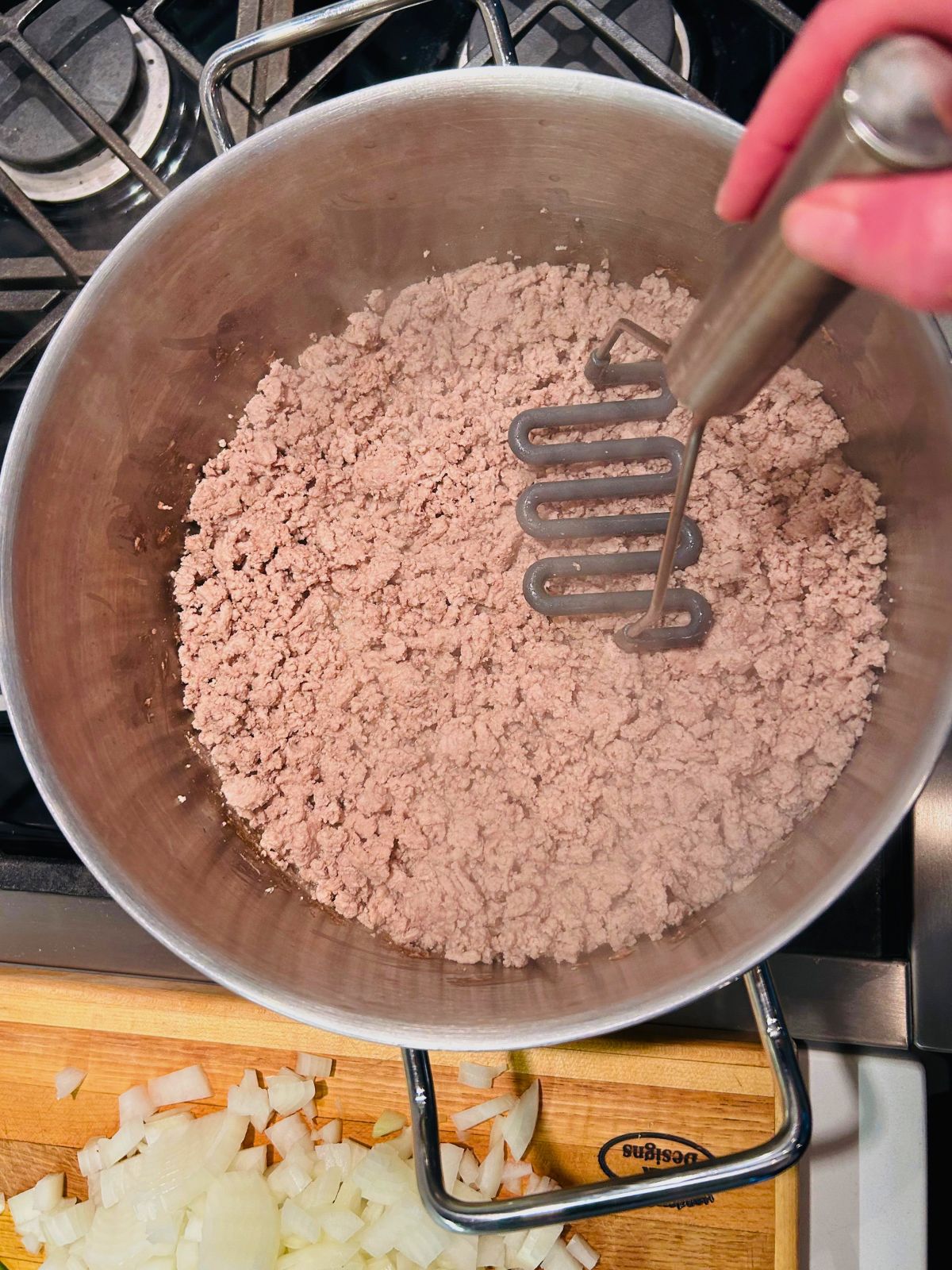 A potato masher is pressed down on the cooked crumbled meat to make the pieces smaller.