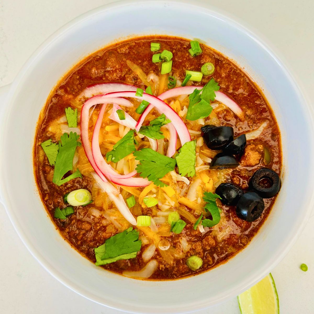 A bowl of thick homemade chili with toppings of onions, cilantro, black olives and cheese.