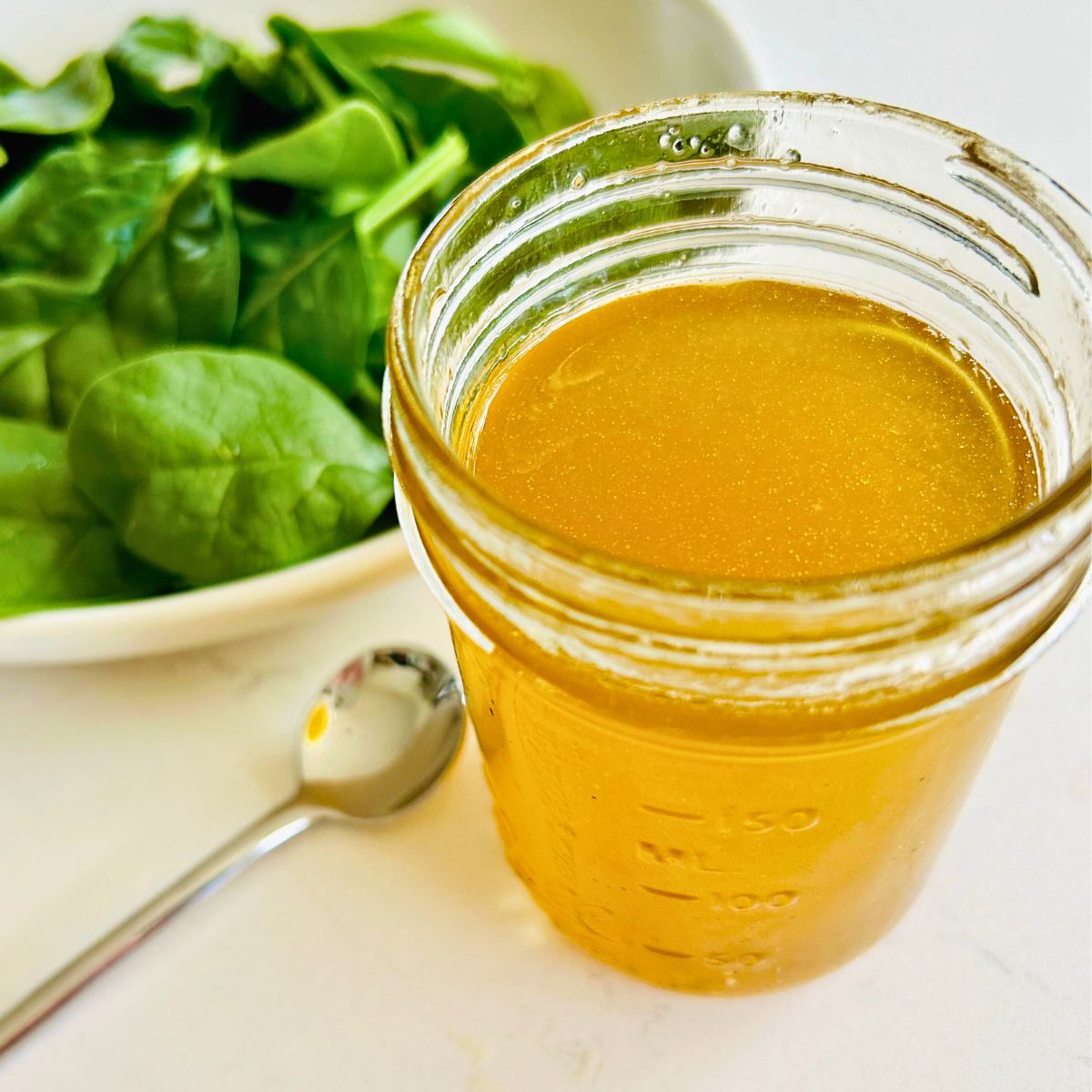 A mason jar of white balsamic vinaigrette next to a bowl of spinach and spoon.