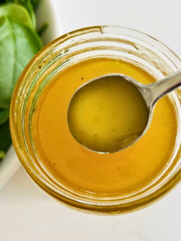 White balsamic vinaigrette in a spoon above a mason jar next to a bowl of spinach leaves.