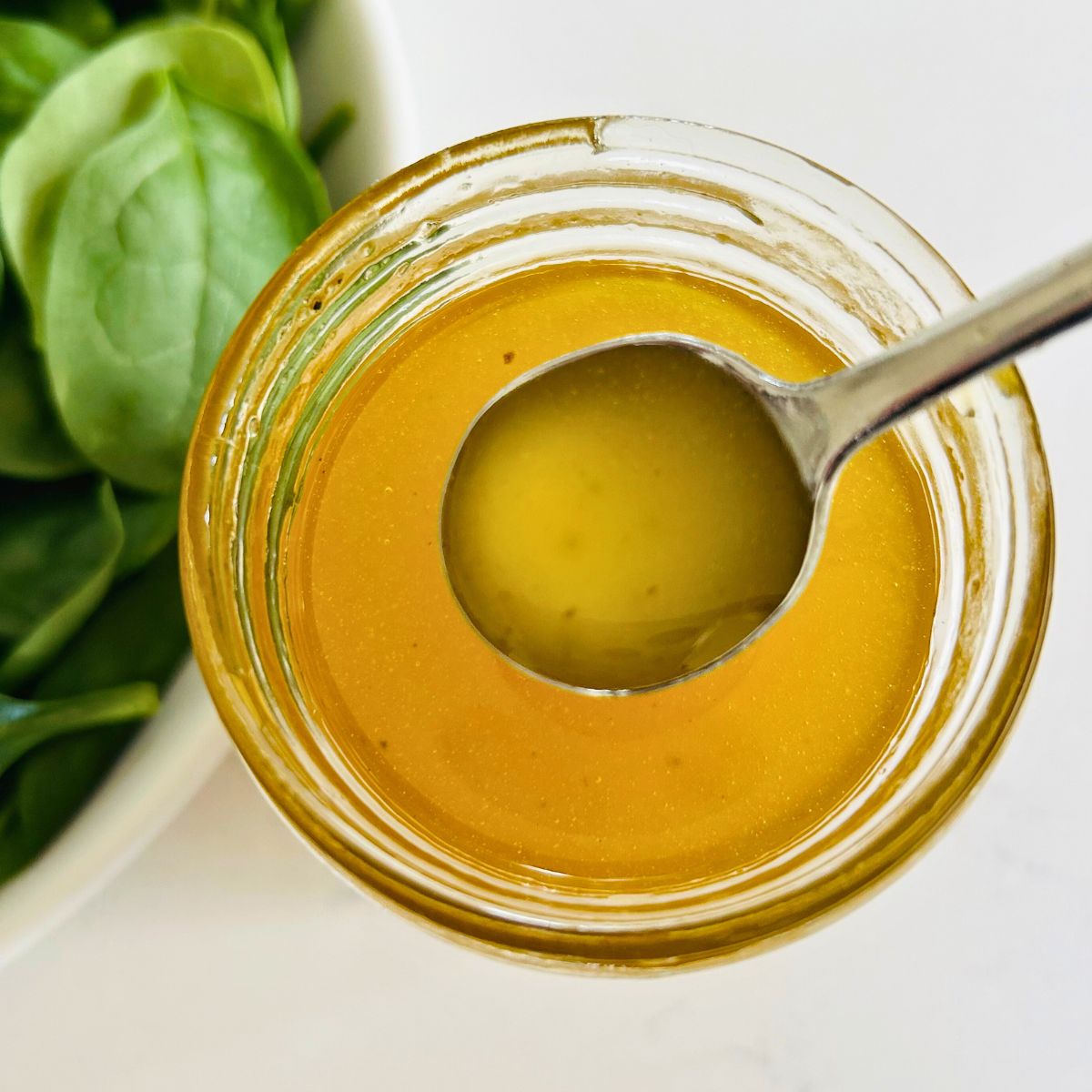 White balsamic vinaigrette in a spoon above a mason jar next to a bowl of spinach leaves.