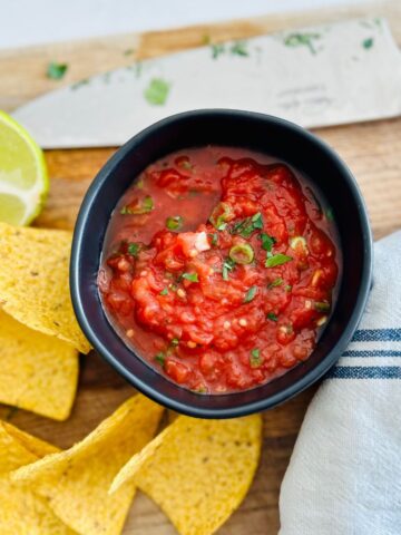 Red restaurant style salsa made from canned tomatoes in a black bowl next to tortilla chips.