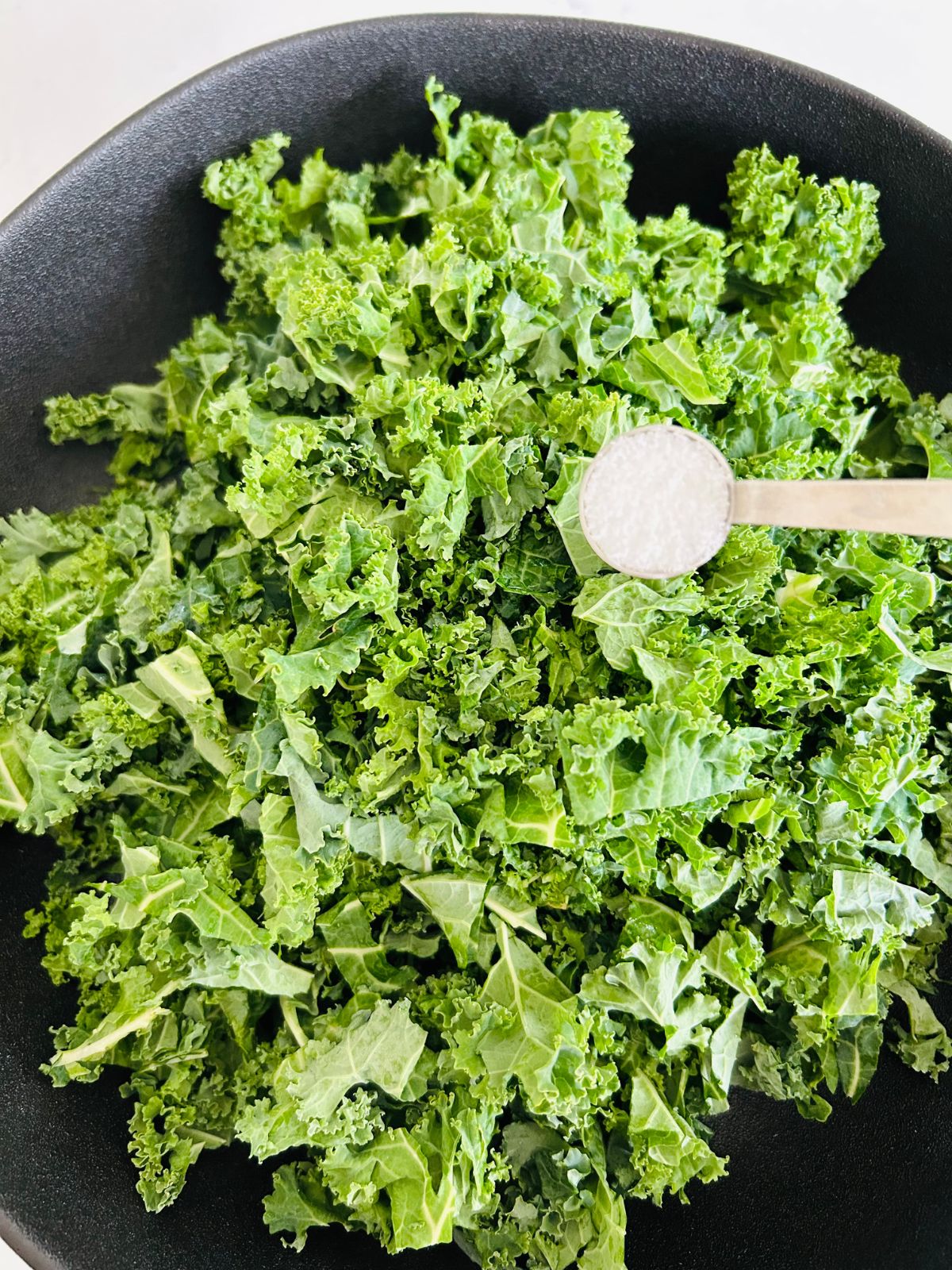 Chopped kale leaves in a salad bowl with a half teaspoon of kosher salt above it.