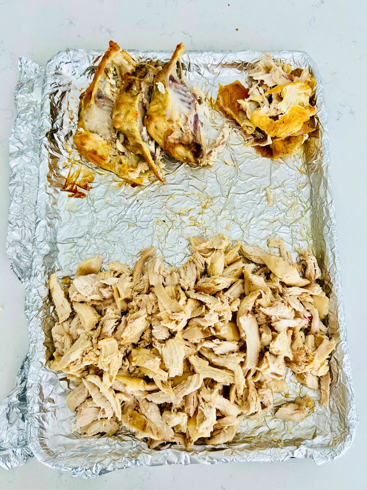 Hand shredded chicken breast with the bones and skin in small piles on a foil lined sheet pan.